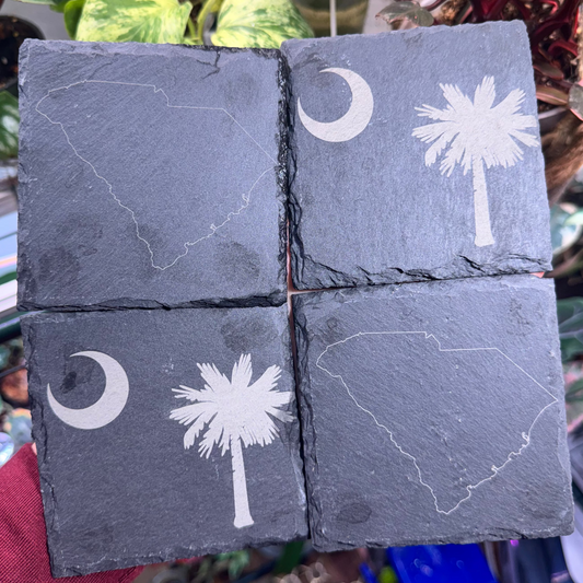 Four slate tiles with white designs of a palm tree, moon, and state outline held in front of plants.